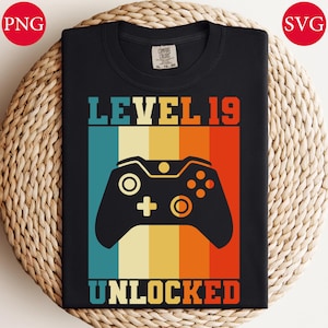 May include: Black t-shirt with a retro design featuring a video game controller and the text "LEVEL 19 UNLOCKED" in a colorful, vertical striped background. The shirt is made of a soft material and is suitable for casual wear.
