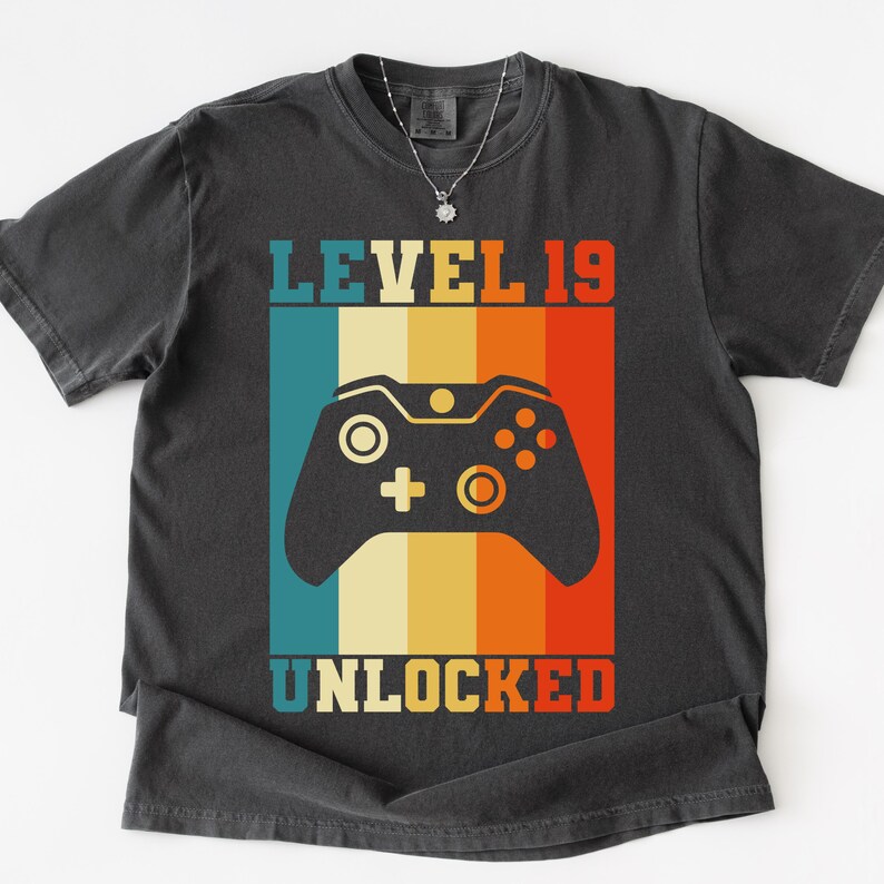 May include: Dark gray t-shirt with a retro design featuring a video game controller and the text "LEVEL 19 UNLOCKED" in a colorful, vertical stripe pattern. The shirt has a crew neck and short sleeves.