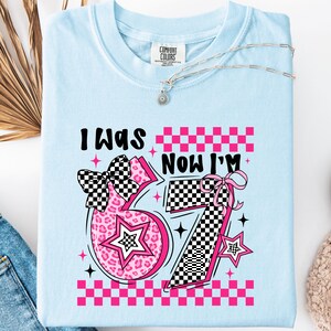 May include: Pale blue t-shirt featuring the text "I WAS... NOW I'M..." and the number "37" in a pink and black checkered design. The shirt has a crew neck and short sleeves. A silver necklace is draped across the shirt.