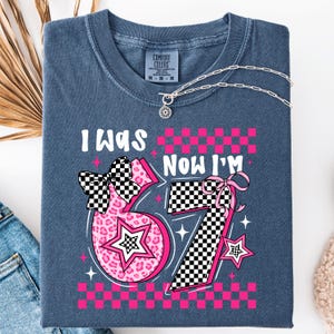 May include: A folded, blue t-shirt with the text "I WAS" above a pink number "67" and "NOW I'M" above a pink number "7". The numbers have a checkered pattern and star accents. A silver necklace is draped across the shirt.