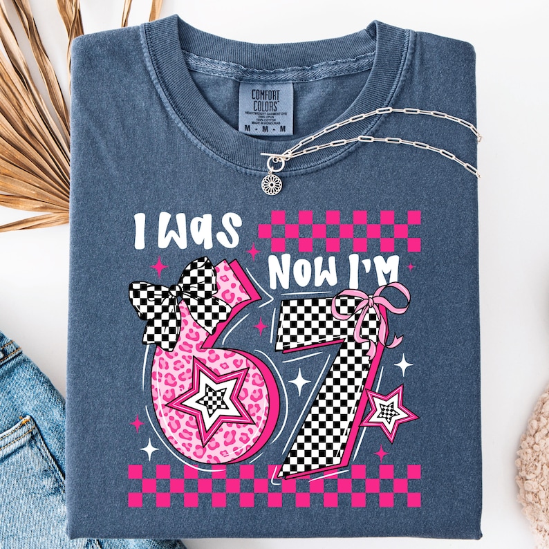 May include: A folded, blue t-shirt with the text "I WAS" above a pink number "67" and "NOW I'M" above a pink number "7". The numbers have a checkered pattern and star accents. A silver necklace is draped across the shirt.