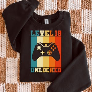 May include: Black sweatshirt with a retro design featuring a video game controller and the text "LEVEL 19 UNLOCKED" in a colorful, striped background. The design is in shades of blue, orange, yellow, and red.