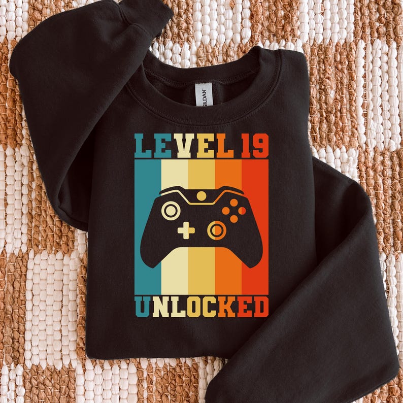 May include: Black sweatshirt with a retro design featuring a video game controller and the text "LEVEL 19 UNLOCKED" in a colorful, striped background. The design is in shades of blue, orange, yellow, and red.