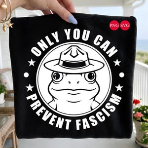 Könnte beinhalten: Schwarzes T-Shirt mit einer weißen Grafik einer Frosch mit Rangerhut, umgeben von dem Text "ONLY YOU CAN PREVENT FASCISM" und Sternen. Das Design enthält "PNG SVG" in roten Kreisen.