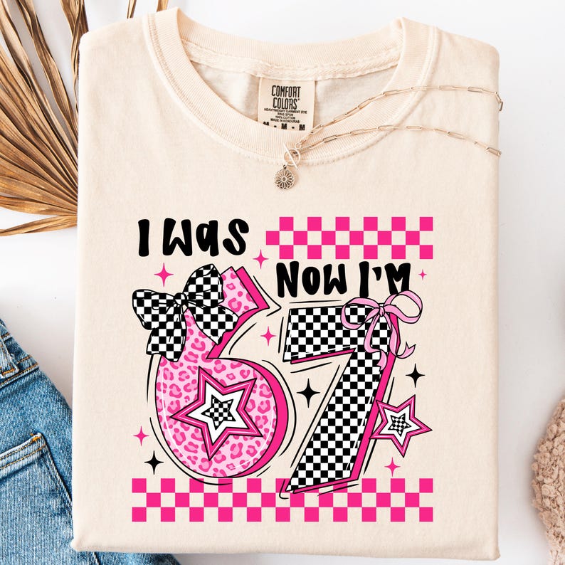 May include: A cream-coloured t-shirt featuring the text "I WAS NOW I'M" above a large pink number "37". The number has a checkered pattern and a star design. A black and white checkered bow is on the left side of the number.