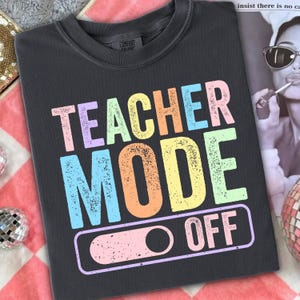 May include: Dark gray t-shirt with the words "TEACHER MODE OFF" in pastel colors. The text is in a bold, playful font. The shirt has a crew neck and a relaxed fit, perfect for casual wear.