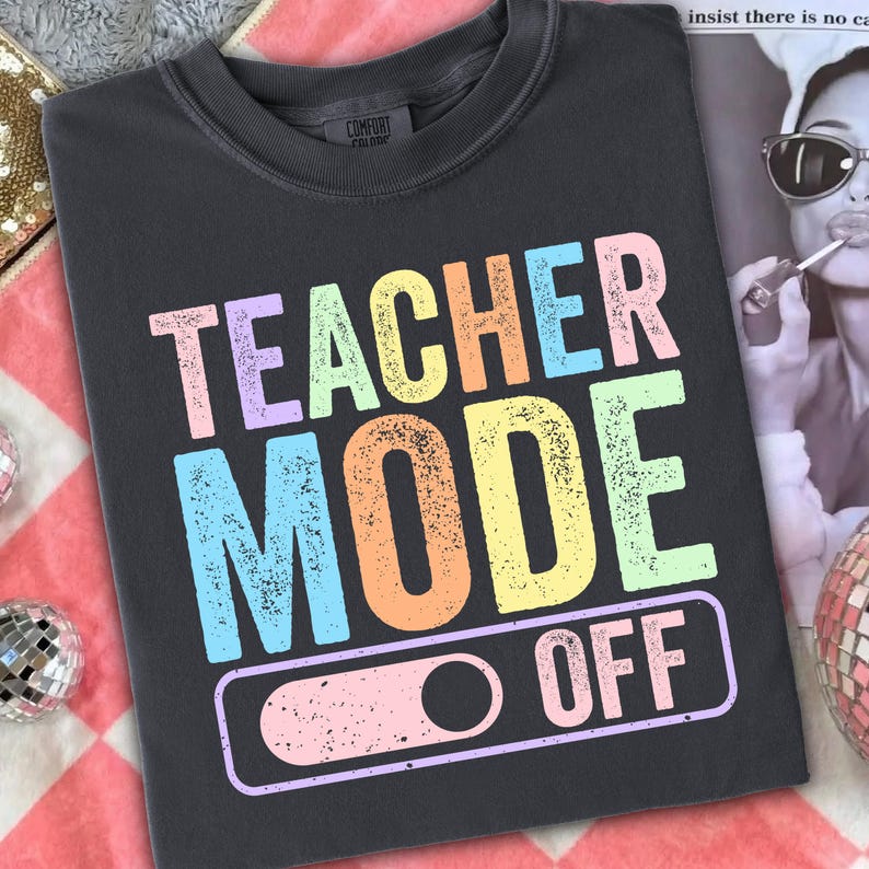 May include: Dark gray t-shirt with the words "TEACHER MODE OFF" in pastel colors. The text is in a bold, playful font. The shirt has a crew neck and a relaxed fit, perfect for casual wear.