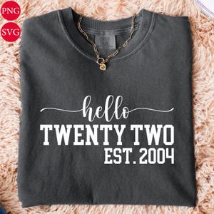 Könnte beinhalten: Ein dunkelgraues T-Shirt mit dem weißen Schriftzug "hello TWENTY TWO EST. 2004". Eine goldene Halskette mit Anhänger liegt auf dem Shirt. Das Shirt liegt auf einer strukturierten, hellbraunen Oberfläche.
