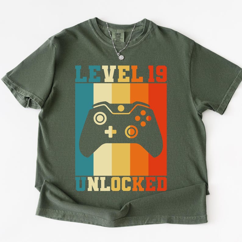 May include: Olive green t-shirt with a retro design. The graphic features a game controller and the text "LEVEL 19 UNLOCKED" in a colorful, vertical striped pattern. The shirt has a crew neck and short sleeves.