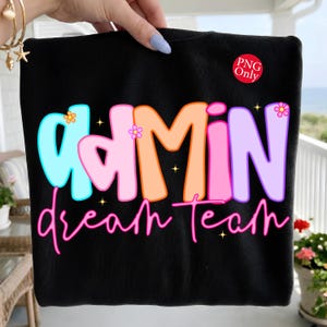 May include: A black sweatshirt featuring the text "adMIN" in large, colourful, bubble-style letters in shades of blue, pink, orange, and purple, with the words "dream Team" in a bright pink script font below. Small flower and star icons are included.