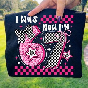 May include: Black t-shirt with the text "I WAS NOW I'M" and the number 67 in pink and white. The number 6 has a leopard print design, and both numbers have star accents and checkered bows.