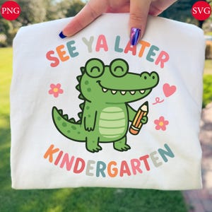 May include: White t-shirt with a colorful graphic of a green alligator wearing glasses and holding a pencil. The text on the shirt reads "SEE YA LATER KINDERGARTEN" in various colors. Pink flowers and a pink heart are also included in the design.