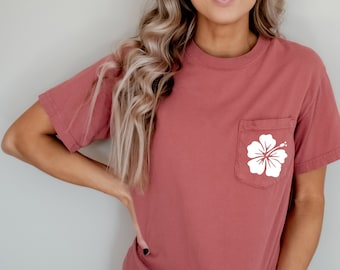 May include: A rust-colored t-shirt with a white hibiscus flower design on the pocket. The text "Cumin" is printed on the bottom right corner of the image.