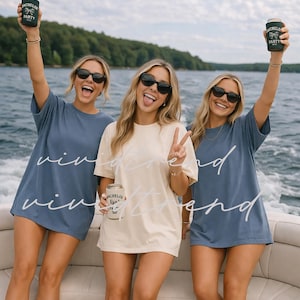May include: Three people wearing sunglasses and t-shirts pose on a boat. Two wear blue shirts with the text "vivid weekend", and one wears a cream-colored shirt. They hold drinks and koozies that say "Bachelor Party".
