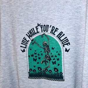 May include: A grey t-shirt with a green and black graphic print of a skeleton dancing in a field of flowers and butterflies. The text "Live While You're Alive" is written in a circle around the skeleton.