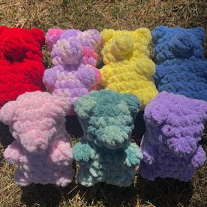 May include: A collection of handmade, plush teddy bears in various colors, including red, pink, yellow, blue, purple, and green. The bears are made of a soft, textured material and are arranged on a grassy surface.