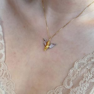 以下が含まれることがあります： 小さな天使のペンダントが付いたゴールドチェーンネックレス。天使はゴールドのドレスを着ており、羽根は一部ゴールド、一部シルバーです。