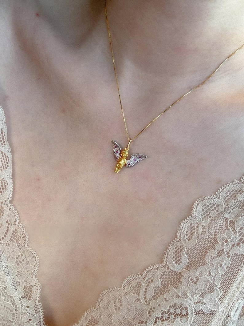 以下が含まれることがあります： 小さな天使のペンダントが付いたゴールドチェーンネックレス。天使はゴールドのドレスを着ており、羽根は一部ゴールド、一部シルバーです。