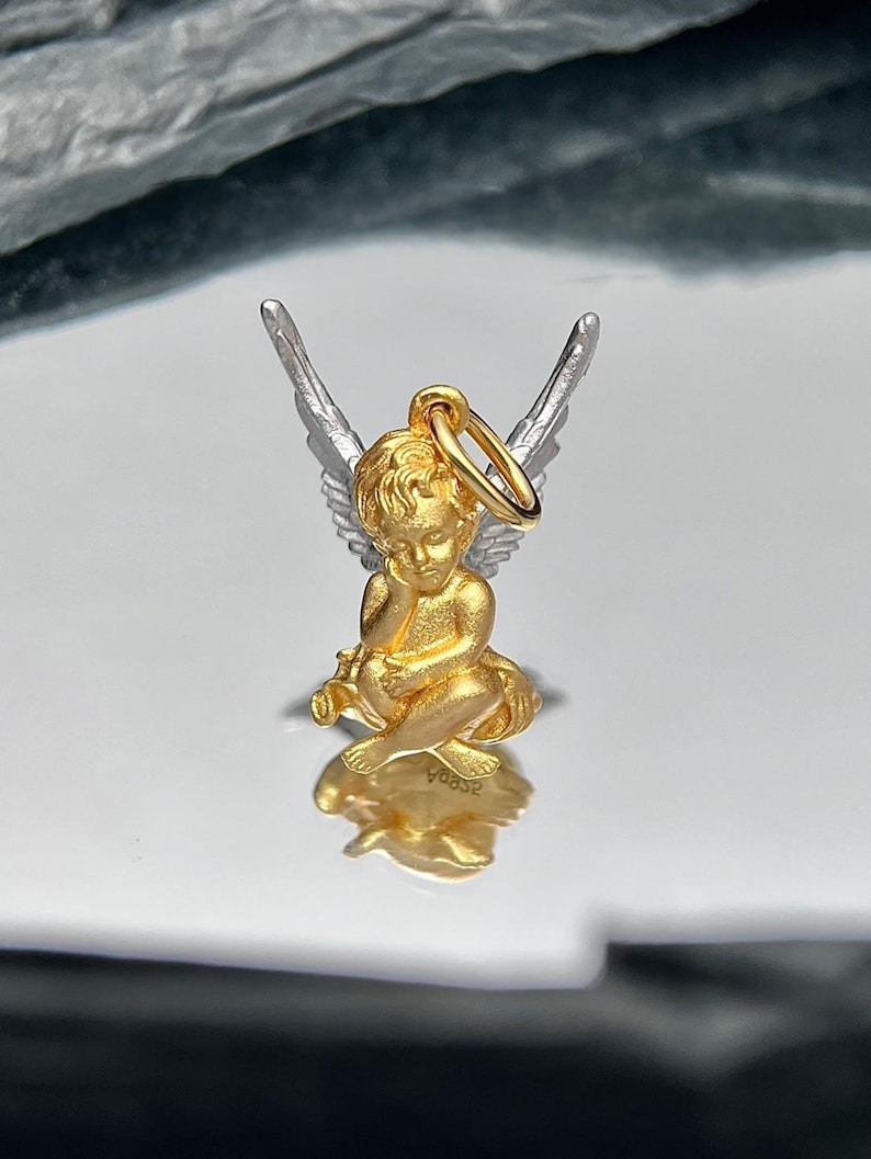 K&ouml;nnte beinhalten: Ein goldener Cherub-Anh&auml;nger mit silbernen Fl&uuml;geln. Der Cherub sitzt mit gefalteten H&auml;nden und hat einen Heiligenschein. Der Anh&auml;nger hat oben eine Schlaufe zum Anbringen an einer Halskette oder einem Armband.
