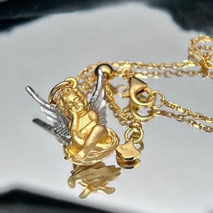 K&ouml;nnte beinhalten: Eine goldene und silberne Halskette mit einem kleinen Amor-Anh&auml;nger. Der Anh&auml;nger ist detailliert mit Fl&uuml;geln verziert und der Amor sitzt mit dem Kopf auf der Hand. Die Halskette hat einen Karabinerverschluss und ein kleines Schild mit der Aufschrift "Ag925".