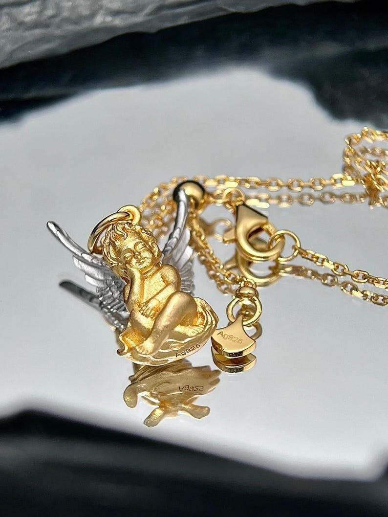 K&ouml;nnte beinhalten: Eine goldene und silberne Halskette mit einem kleinen Amor-Anh&auml;nger. Der Anh&auml;nger ist detailliert mit Fl&uuml;geln verziert und der Amor sitzt mit dem Kopf auf der Hand. Die Halskette hat einen Karabinerverschluss und ein kleines Schild mit der Aufschrift "Ag925".