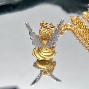 K&ouml;nnte beinhalten: Goldener und silberner Engel-Anh&auml;nger mit ausgebreiteten Fl&uuml;geln. Der Anh&auml;nger befindet sich an einer goldenen Kette.