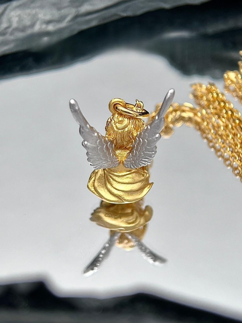 K&ouml;nnte beinhalten: Goldener und silberner Engel-Anh&auml;nger mit ausgebreiteten Fl&uuml;geln. Der Anh&auml;nger befindet sich an einer goldenen Kette.