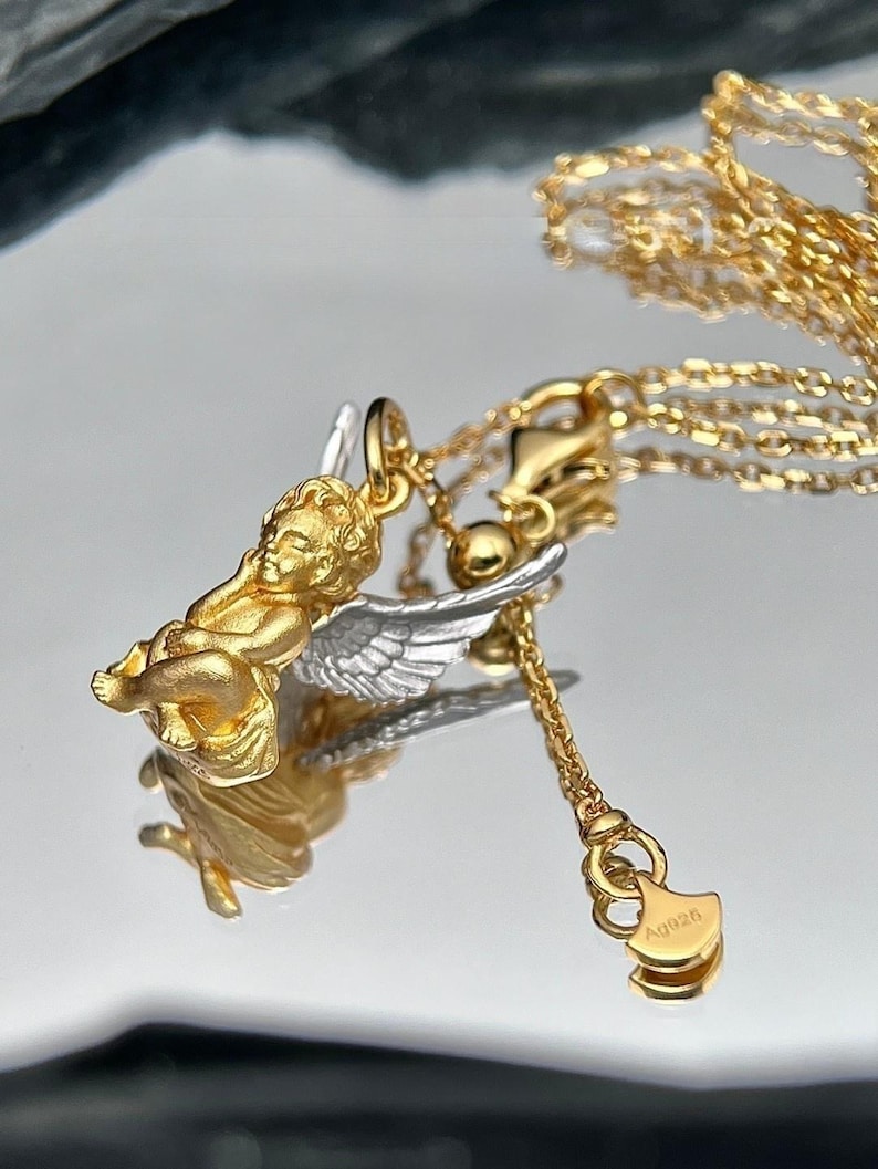 K&ouml;nnte beinhalten: Goldene und silberne Halskette mit einem Amor-Anh&auml;nger. Der Amor sitzt mit ausgebreiteten Fl&uuml;geln. Die Halskette hat einen kleinen Anh&auml;nger mit dem Text "AQ926".