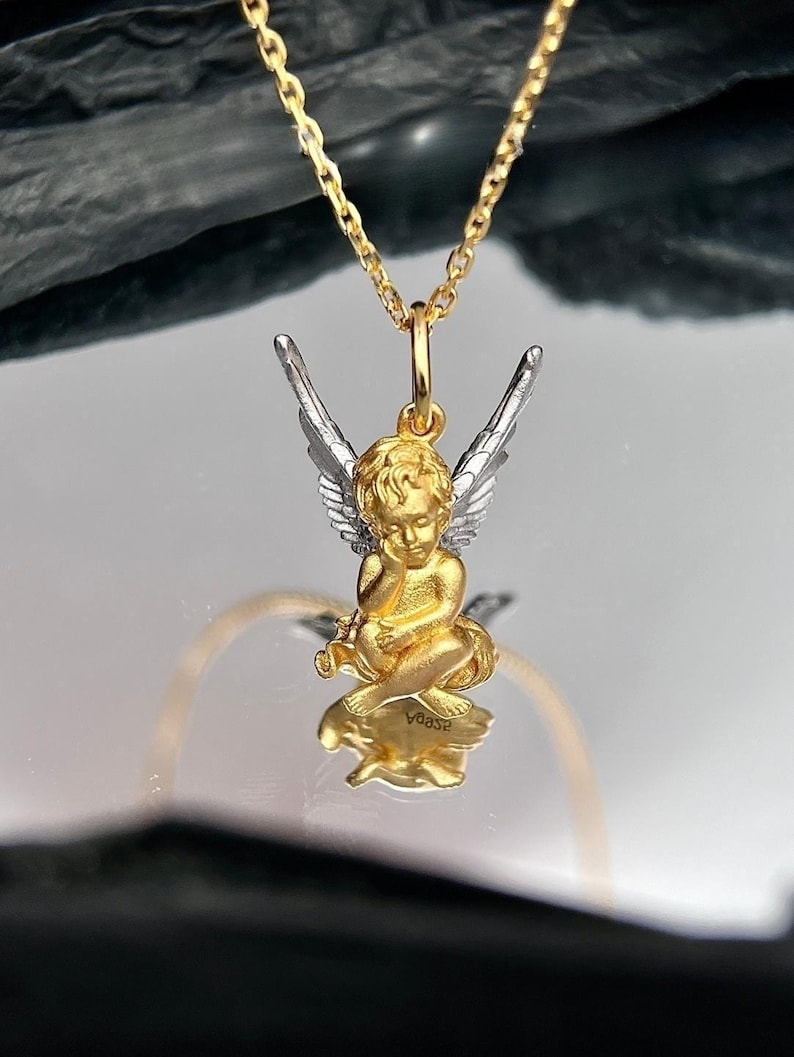 K&ouml;nnte beinhalten: Eine Halskette mit einem goldenen und silbernen Engel-Anh&auml;nger. Der Engel sitzt mit ausgebreiteten Fl&uuml;geln. Der Anh&auml;nger ist an einer goldenen Kette.