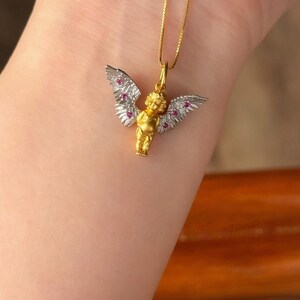 以下が含まれることがあります： 白い羽根とピンクの宝石が付いた金の天使のペンダント。ペンダントは金のチェーンに付いています。