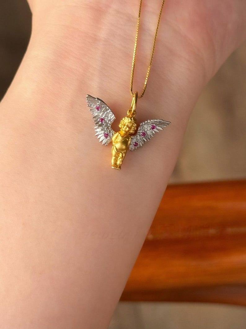 以下が含まれることがあります： 白い羽根とピンクの宝石が付いた金の天使のペンダント。ペンダントは金のチェーンに付いています。