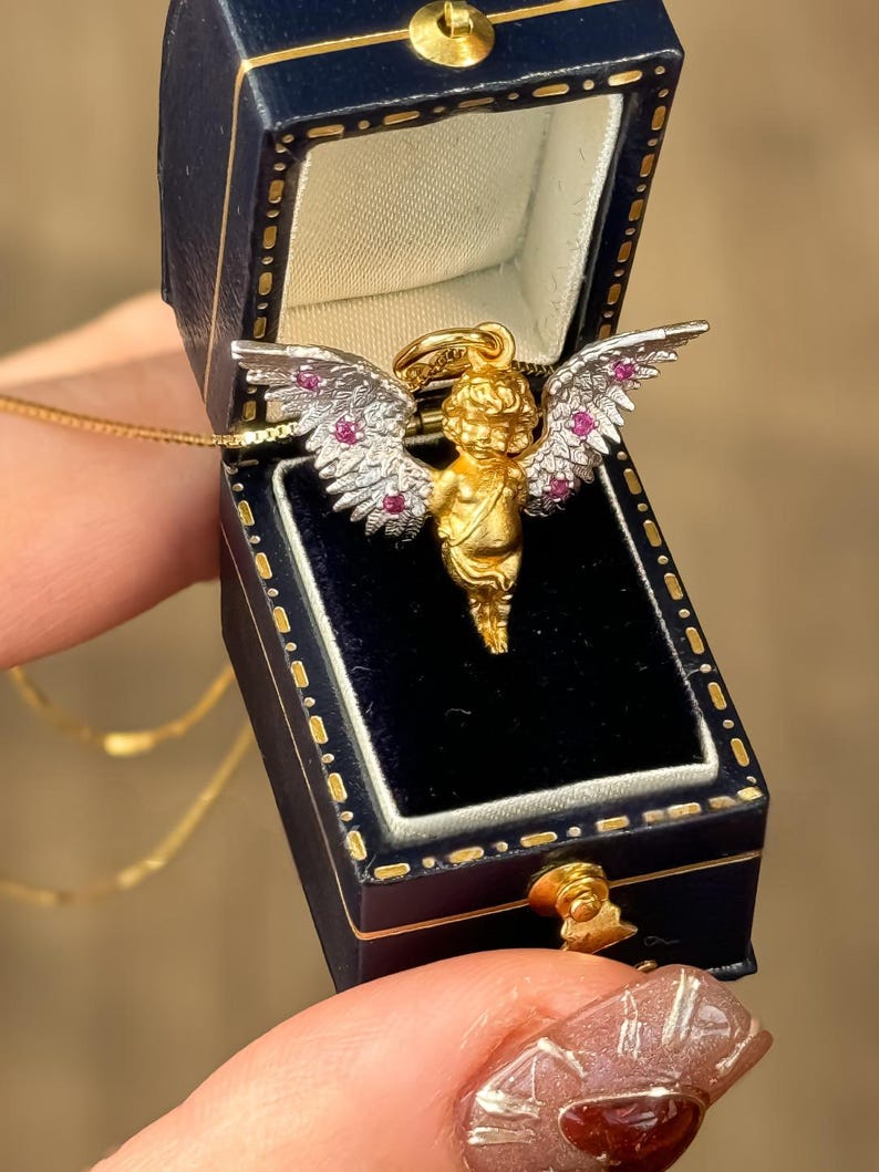 以下が含まれることがあります： 青いベルベットのジュエリーボックスに入った、ルビーのアクセントと白い羽根を持つ金のケルビムペンダント