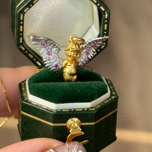 以下が含まれることがあります： ピンクと白の羽根を持つ金のキューピッドペンダント。小さなピンクの宝石がセットされています。ペンダントは緑色のベルベット製のリングボックスに入っています。
