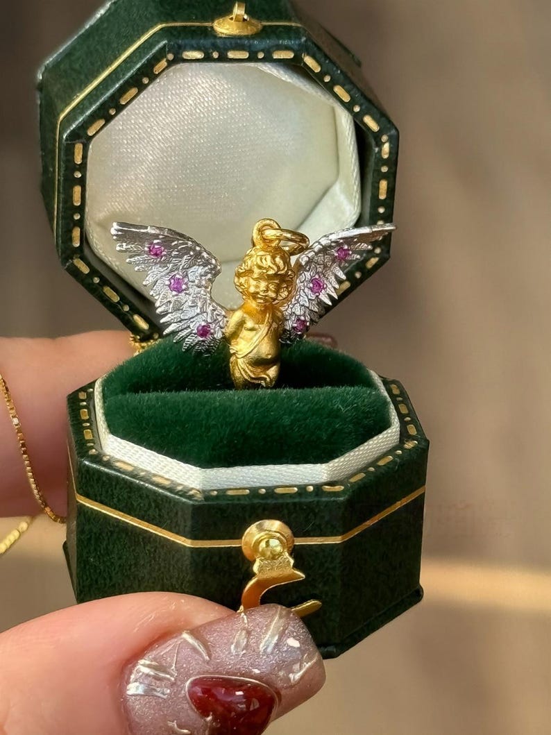 以下が含まれることがあります： ピンクと白の羽根を持つ金のキューピッドペンダント。小さなピンクの宝石がセットされています。ペンダントは緑色のベルベット製のリングボックスに入っています。