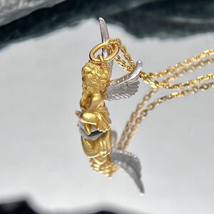 K&ouml;nnte beinhalten: Ein goldener Cherub-Anh&auml;nger mit silbernen Fl&uuml;geln h&auml;ngt an einer goldenen Kette. Der Anh&auml;nger ist detailliert und hat ein realistisches Cherub-Design.