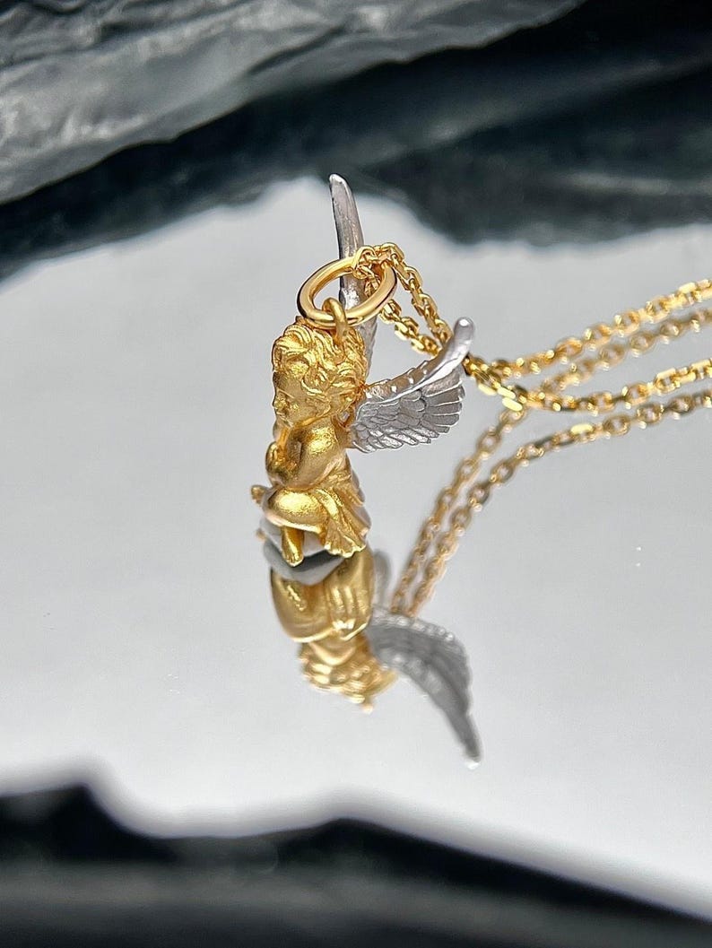 K&ouml;nnte beinhalten: Ein goldener Cherub-Anh&auml;nger mit silbernen Fl&uuml;geln h&auml;ngt an einer goldenen Kette. Der Anh&auml;nger ist detailliert und hat ein realistisches Cherub-Design.