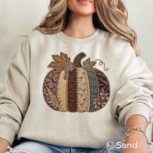May include: A sand-colored crewneck sweatshirt featuring a large, central pumpkin graphic. The pumpkin is composed of vertical fabric panels in shades of brown, tan, and olive, decorated with embroidery, buttons, and floral patterns.