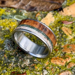May include: A silver ring with a wooden inlay featuring green and white accents. The ring is on a bed of green moss and brown rocks.