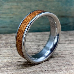 May include: A silver ring with a brown wood inlay. The ring is on a wooden surface.