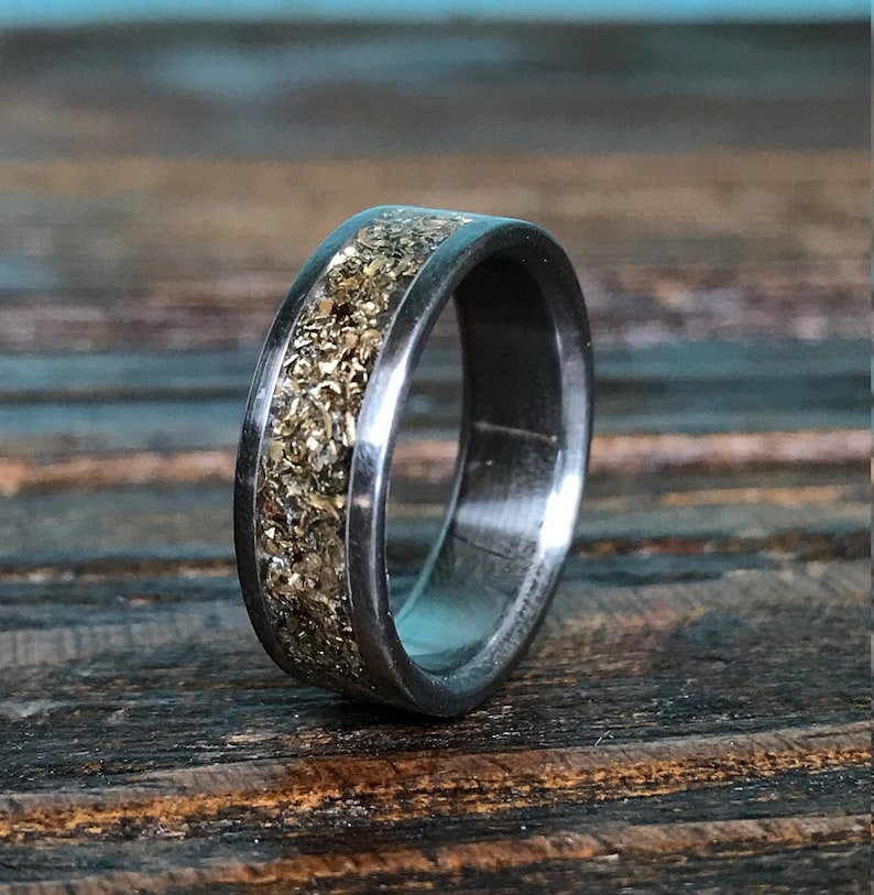 A close-up shot of a dark metallic ring showing a speckled, glittering gold inlay running along its center.