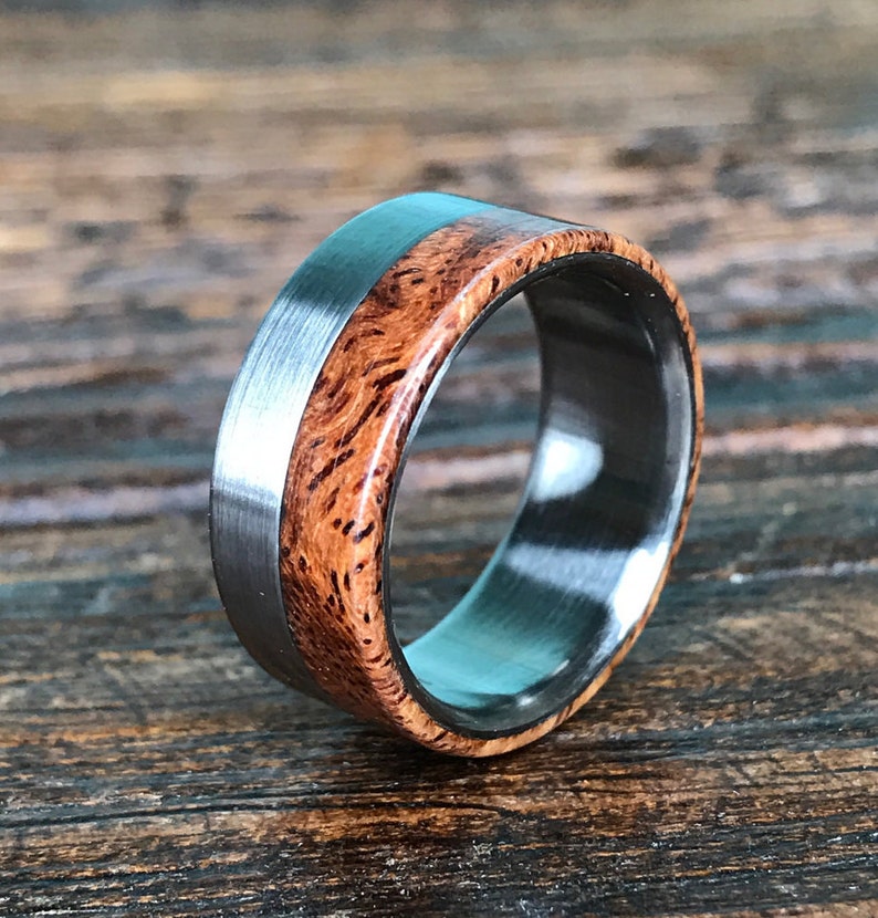 May include: A silver and brown wood ring with a brushed finish. The ring has a wide band with a thin silver strip around the edge.
