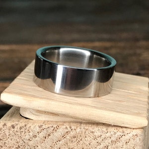 May include: A silver-toned metal ring with a flat, wide band. The ring is sitting on a wooden surface.