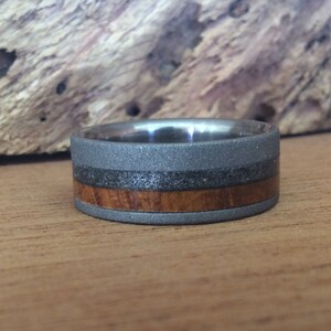 May include: A gray titanium ring with a brown wood inlay and a black band. The ring has a textured finish.