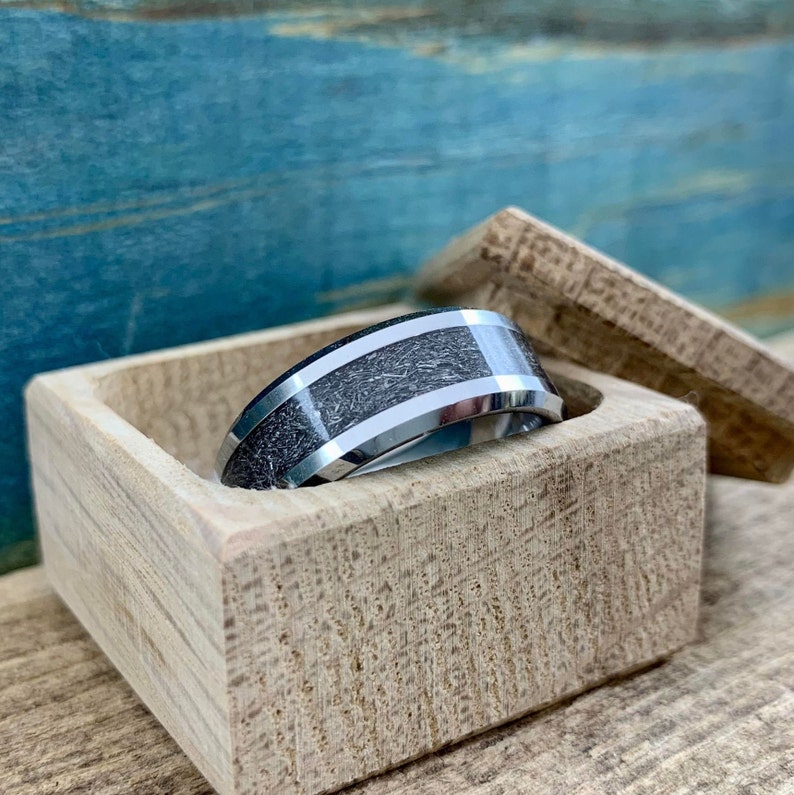 May include: A silver and black wedding band with a wood inlay sits in a wooden ring box.