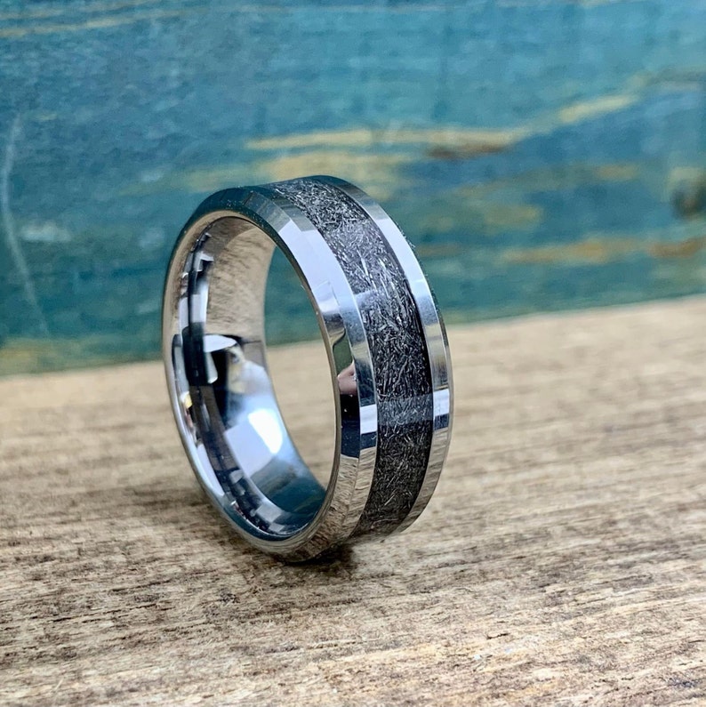 May include: A silver tungsten carbide ring with a beveled edge and a gray inlay. The inlay has a textured, wood-like pattern.