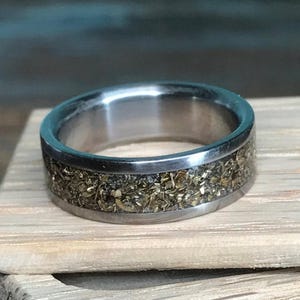 A metallic ring with a textured gold-flake inlay resting upright on a small piece of light-colored wood.