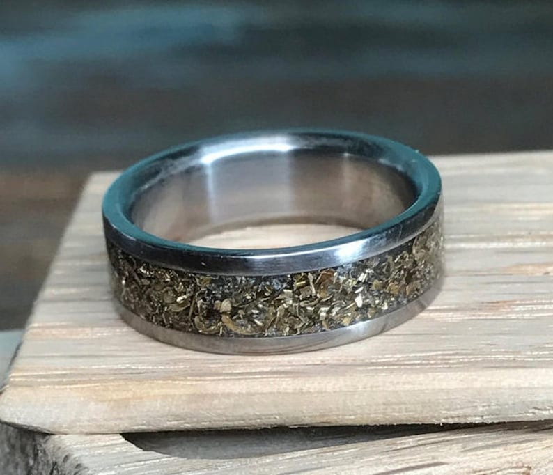 A metallic ring with a textured gold-flake inlay resting upright on a small piece of light-colored wood.