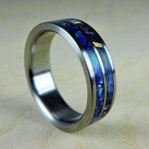 Op de afbeelding: Een metalen ring met een geborstelde zilveren afwerking met een centrale inleg van verbrijzelde blauwe en turquoise steenfragmenten naast donkere houtnerftexturen, wat een ring met meerdere texturen en een glad, gepolijst binnenoppervlak vormt.