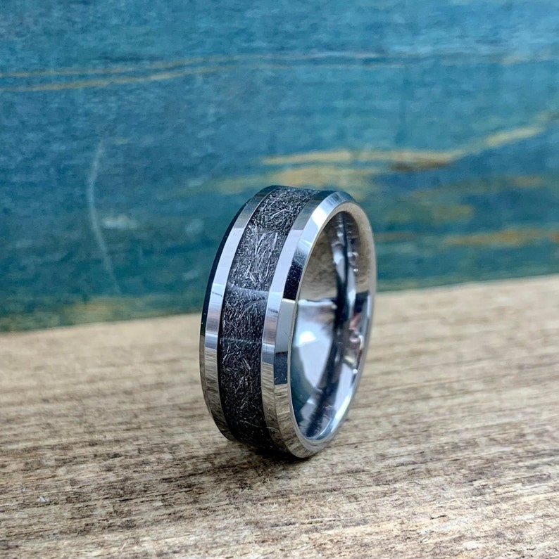 May include: A silver tungsten carbide ring with a beveled edge and a black inlay. The inlay has a wood grain pattern.