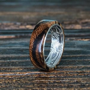 A zebrawood ring with a silver interior and engraved floral pattern is shown from a slightly different angle on a dark wooden surface.